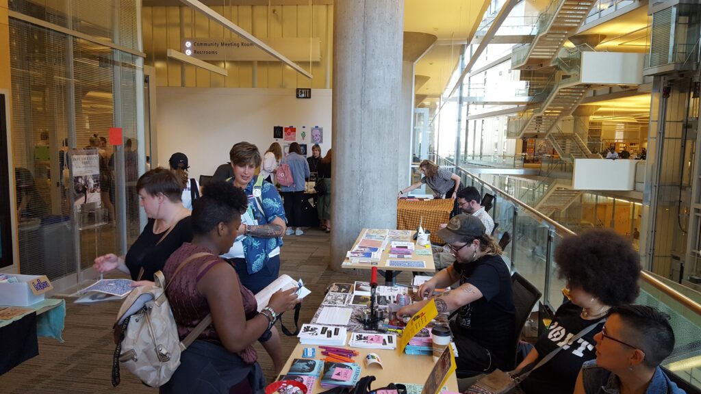 People shop at tables set up in a hallway-type area. There is an open-air atrium behind them with a lot of natural light.